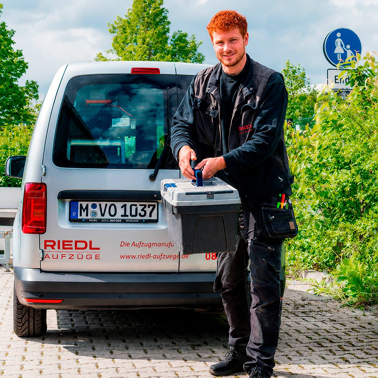 Ein Mitarbeiter steht mit einem Werkzeugkasten vor der Rückseite eines Firmenautos.