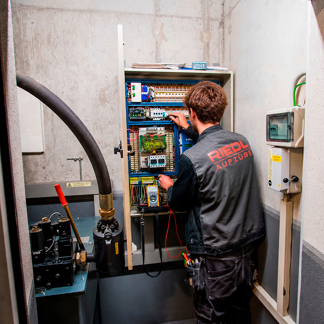 Ein Mitarbeiter steht vor einem Schrank mit Steuerungsmodulen und misst mit einem Multimeter Kontakte durch. Links führt ein dicker Schlauch hinter eine Betonstrebe.